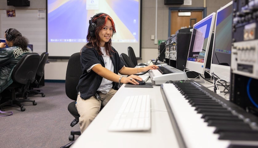 Student wearing headphones works at a computer workstation with audio production equipment and a keyboard piano in a digital media lab, smiling toward the camera.