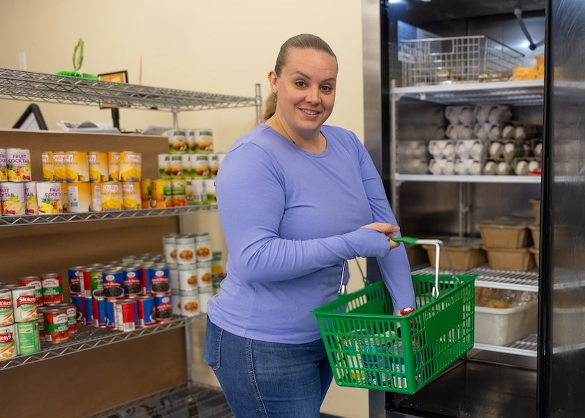 A student smiles while selecting groceries from a campus food pantry, holding a green shopping basket in front of shelves stocked with canned goods, fruit cups, eggs, and refrigerated food.