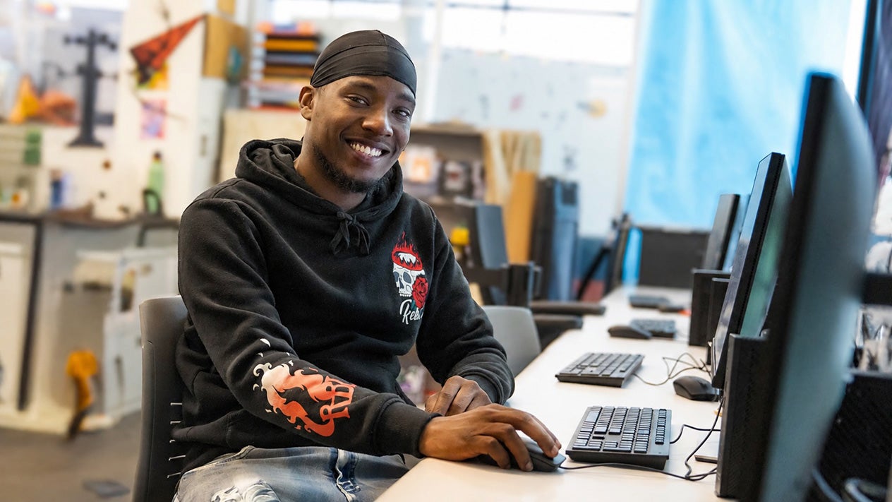 Smiling student wearing a black hoodie and durag sits at a desk using a computer in a bright campus computer lab.