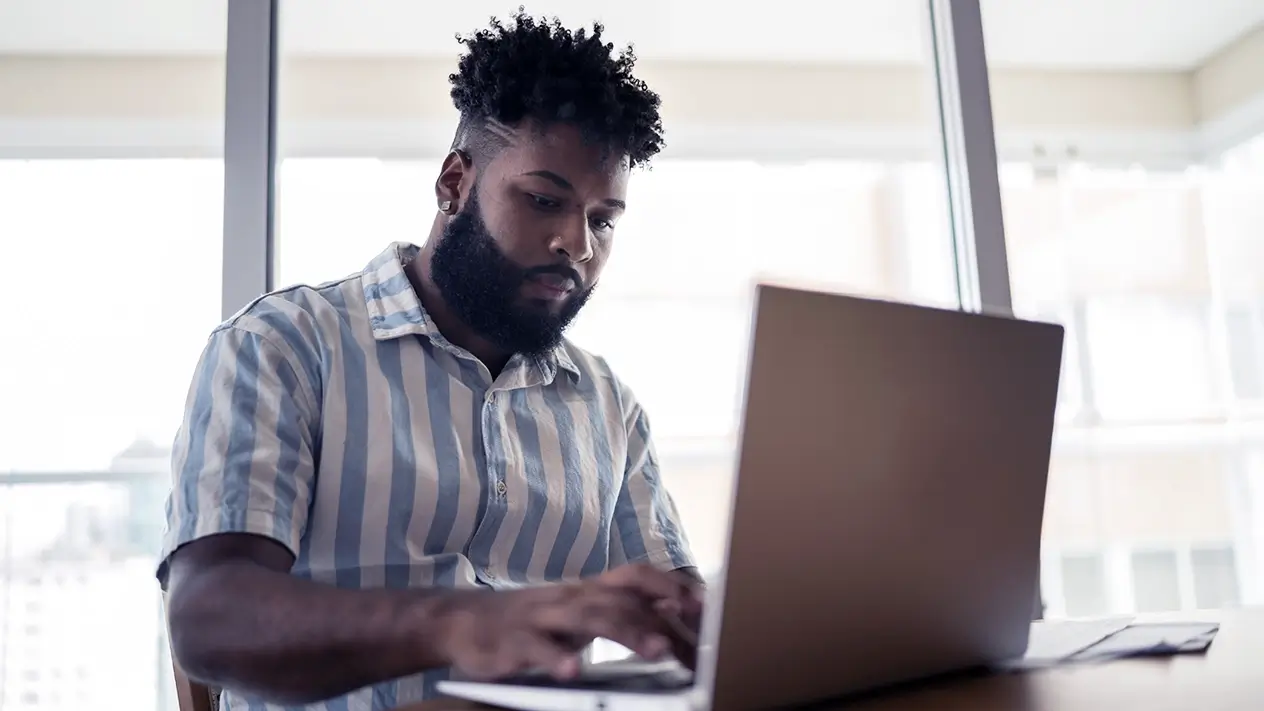 Student sitting at a desk, typing on a laptop, in front of bright window