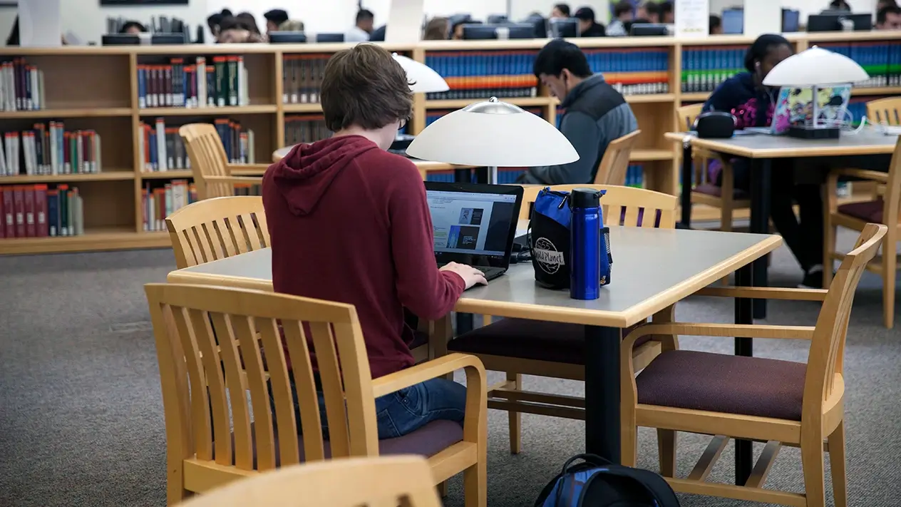 Three students sitting at library tables, surrounded by bookshelf, with more students in background