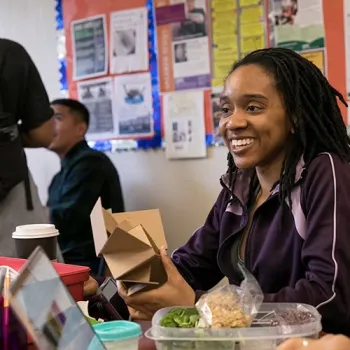 Woman eating healthy vegetables while seated in a cafeteria setting; two students talking in the background.
