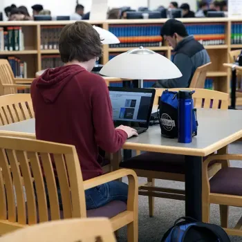 Three students sitting at library tables, surrounded by bookshelf, with more students in background