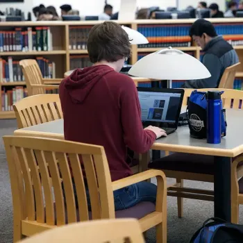 Two students sitting at library tables, surrounded by bookshelf, with more students in background