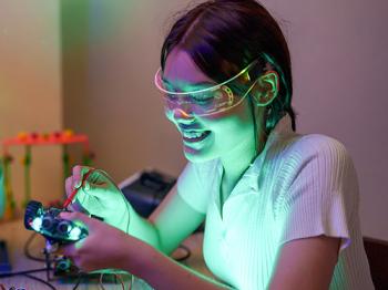 Young woman wearing safety goggles, smiling and focused while working on an electronics project with glowing components on a table.