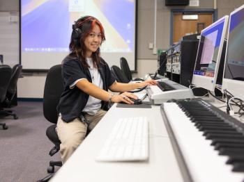 Student wearing headphones works at a computer workstation with audio production equipment and a keyboard piano in a digital media lab, smiling toward the camera.