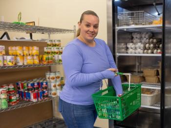 A student smiles while selecting groceries from a campus food pantry, holding a green shopping basket in front of shelves stocked with canned goods, fruit cups, eggs, and refrigerated food.